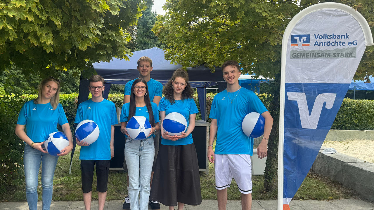 Einige junge Leute in blauem Shirt stehen mit einem blau-weißen Ball in der Hand neben einem Volksbank Aufsteller.