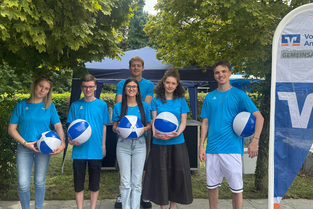 Einige junge Leute in blauem Shirt stehen mit einem blau-weißen Ball in der Hand neben einem Volksbank Aufsteller.