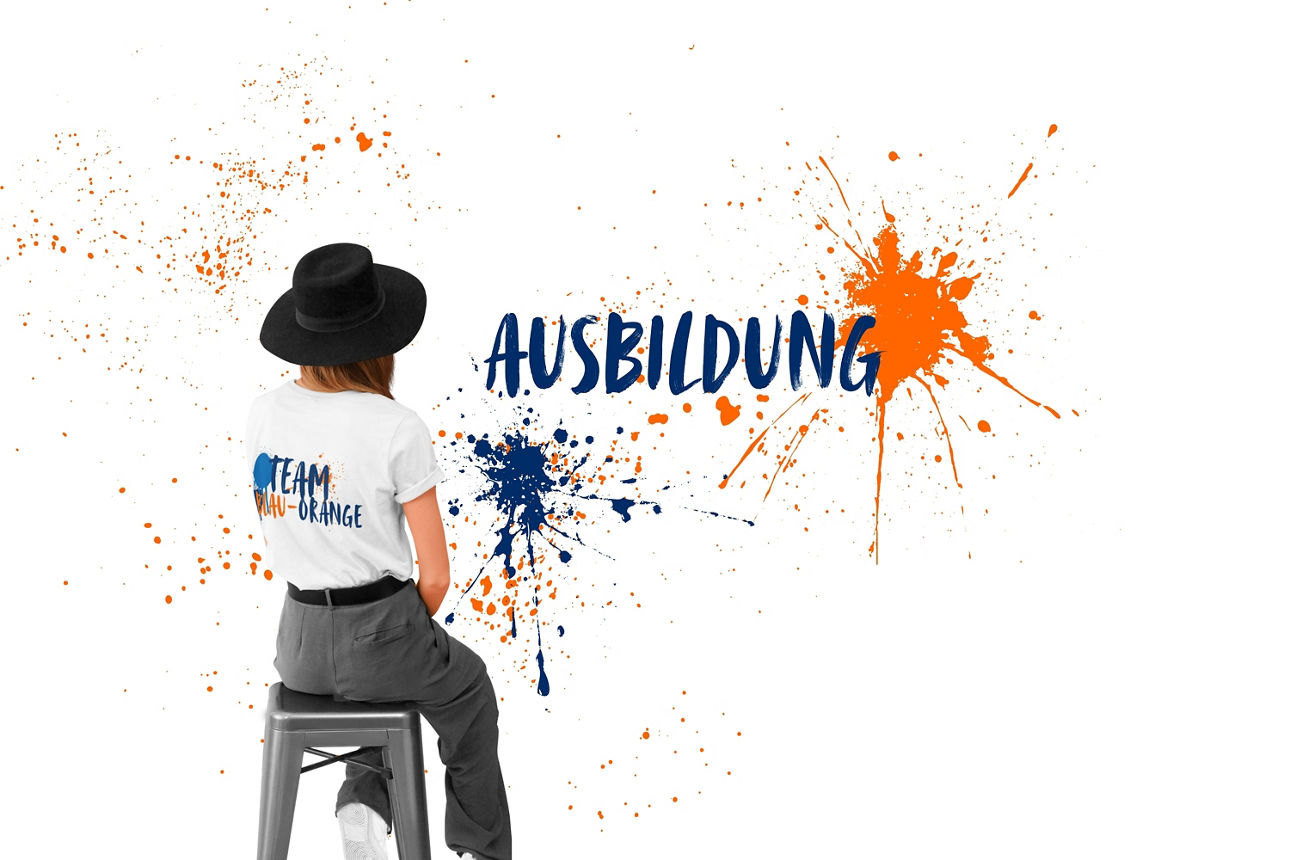 Rothaarige Frau auf Stuhl, trägt „Team Blau-Orange“-T-Shirt, schwarzen Hut, vor weißem Hintergrund mit blauer Aufschrift.