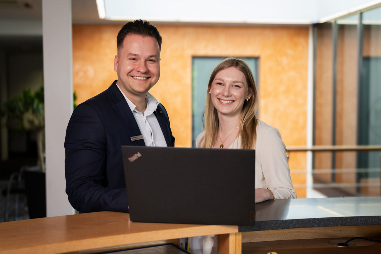 Mann und Frau stehen an einem Bürotresen mit geöffnetem Laptop und lächeln in die Kamera