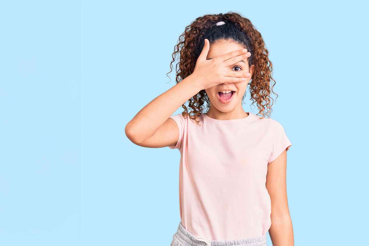 Mädchen schaut fröhlich durch ihre Finger, vor einem hellblauem Hintergrund.