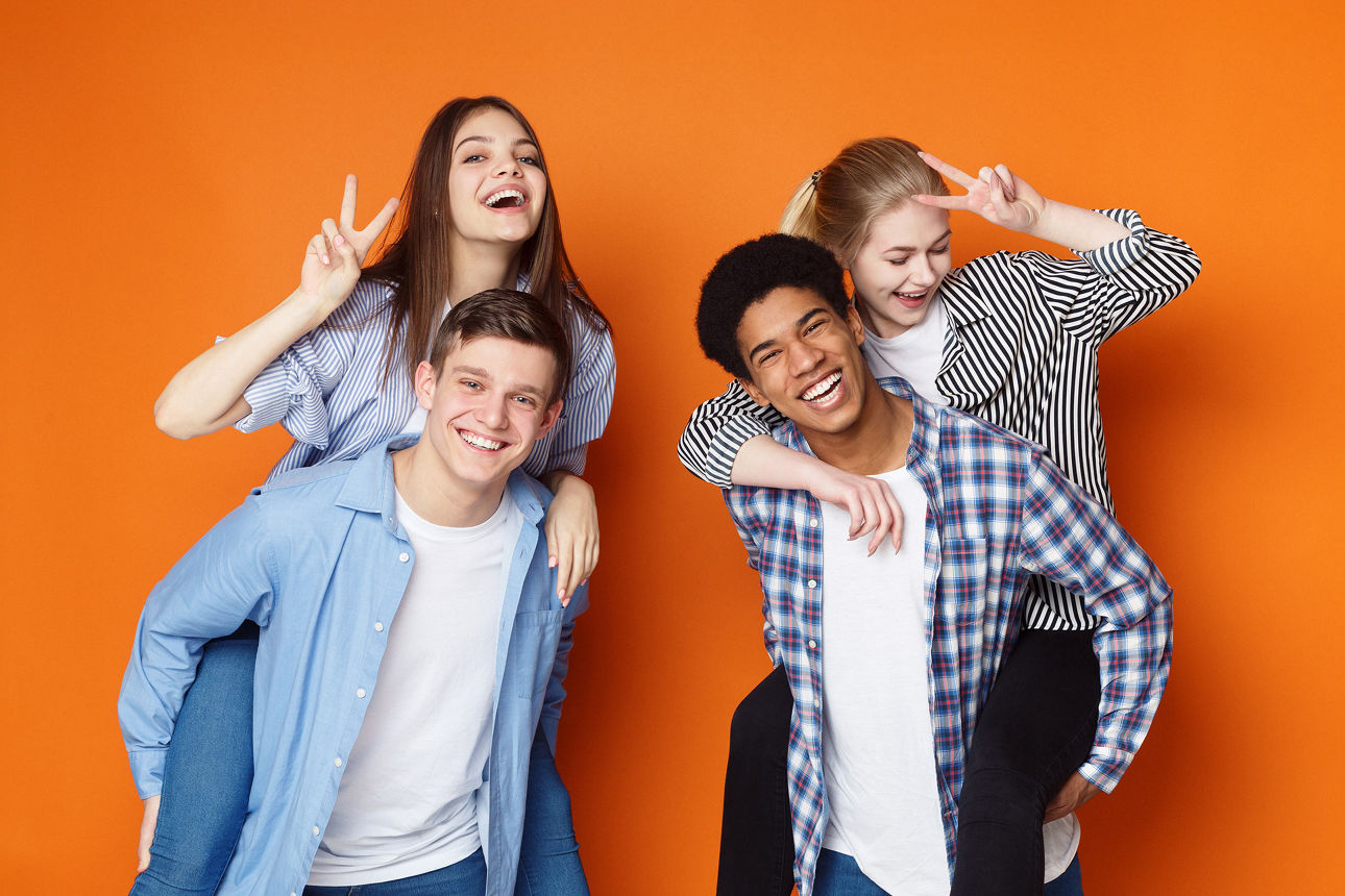 Zwei junge Männer tragen jeweils zwei Junge Frauen huckepack. Die Gruppe steht vor einem orangenem Hintergrund und lächelt in die Kamera.