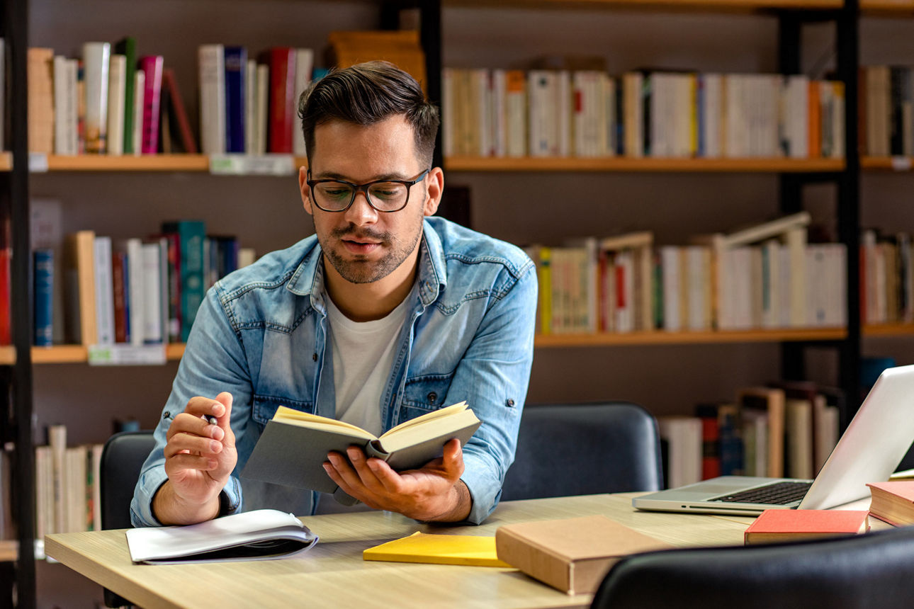 Junger Mann sitzt in der Bibliothek und liest in einem Buch. Um ihn herum liegen Blöcke und ein aufgeklappter Laptop.