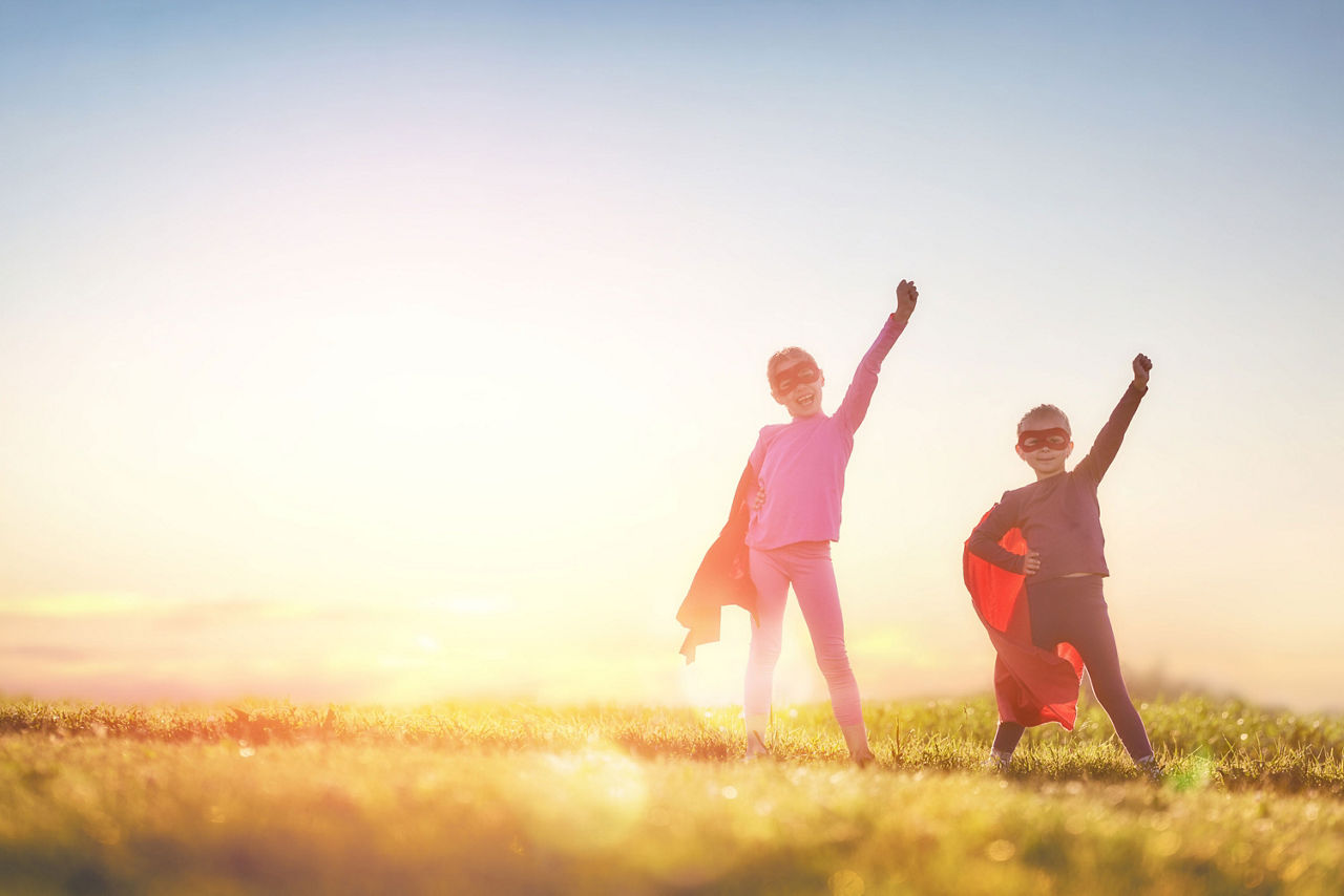 Zwei Kinder mit Superheldenkostümen stehen im Sonnenuntergang und recken die Arme in Superhelden-Pose in die Luft