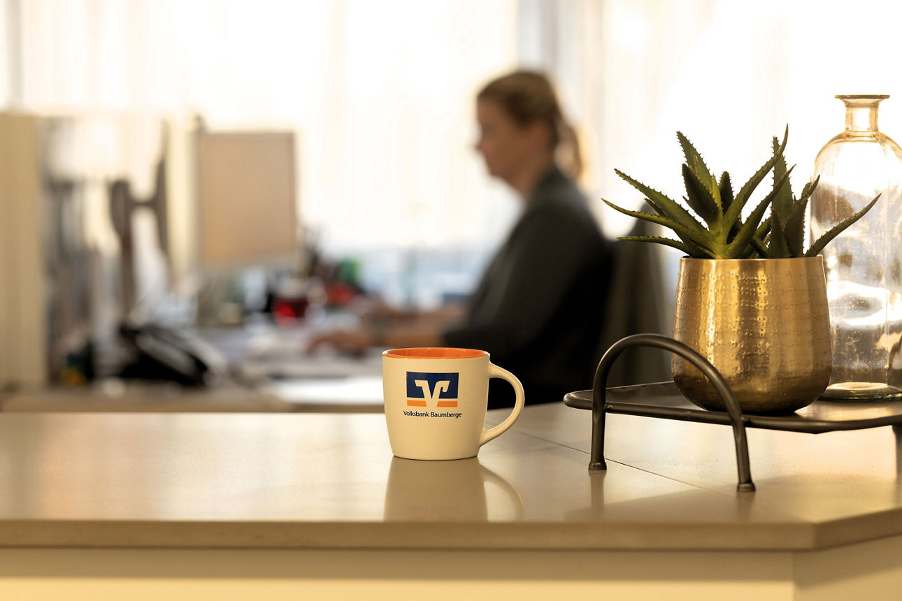 Auf einer Theke steht eine Tasse mit Volksbank‑Logo neben einer Pflanze. Im Hintergrund sitzt eine weibliche Person am PC.