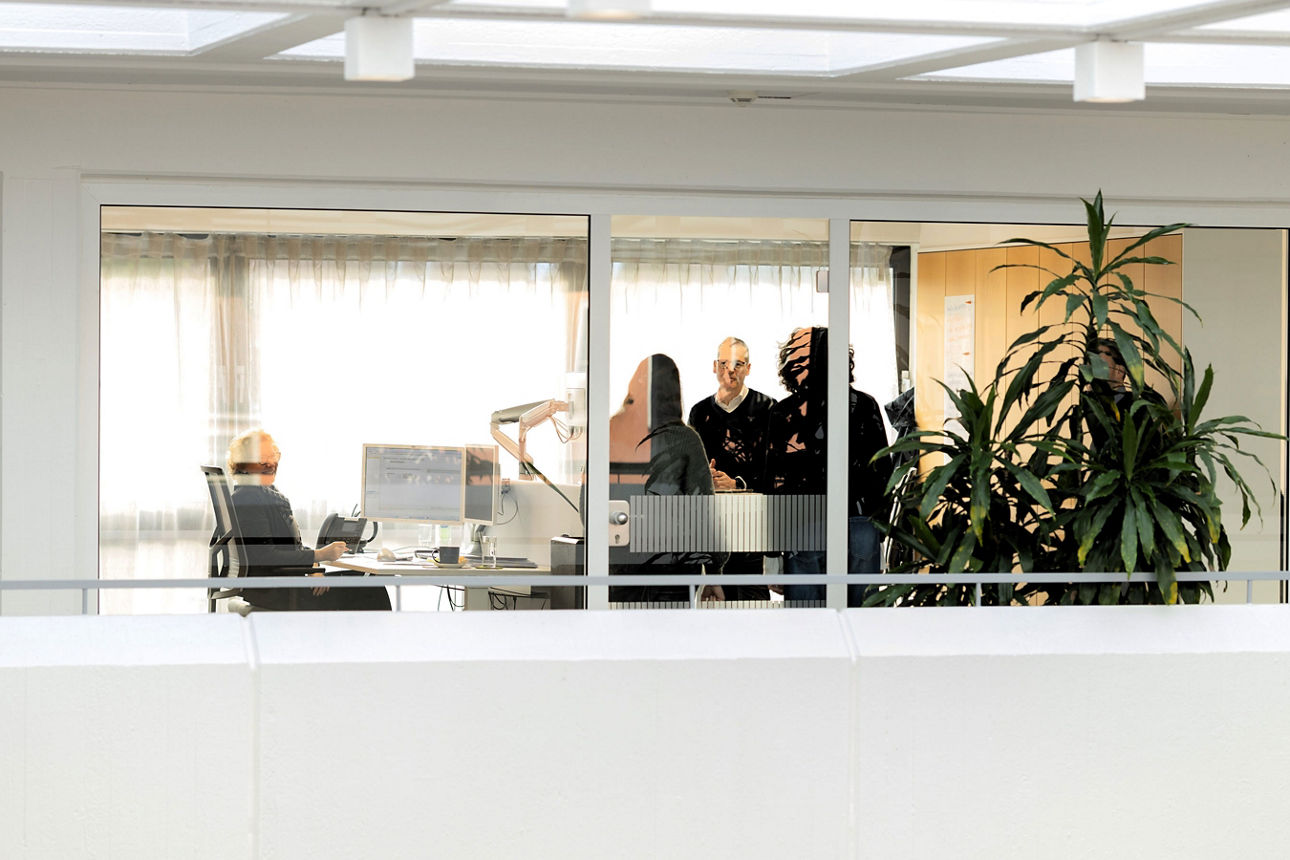 Durch ein Fenster ist ein heller Büroraum mit mehreren Personen, Schreibtischen, Computern und einer großen Pflanze zu sehen.
