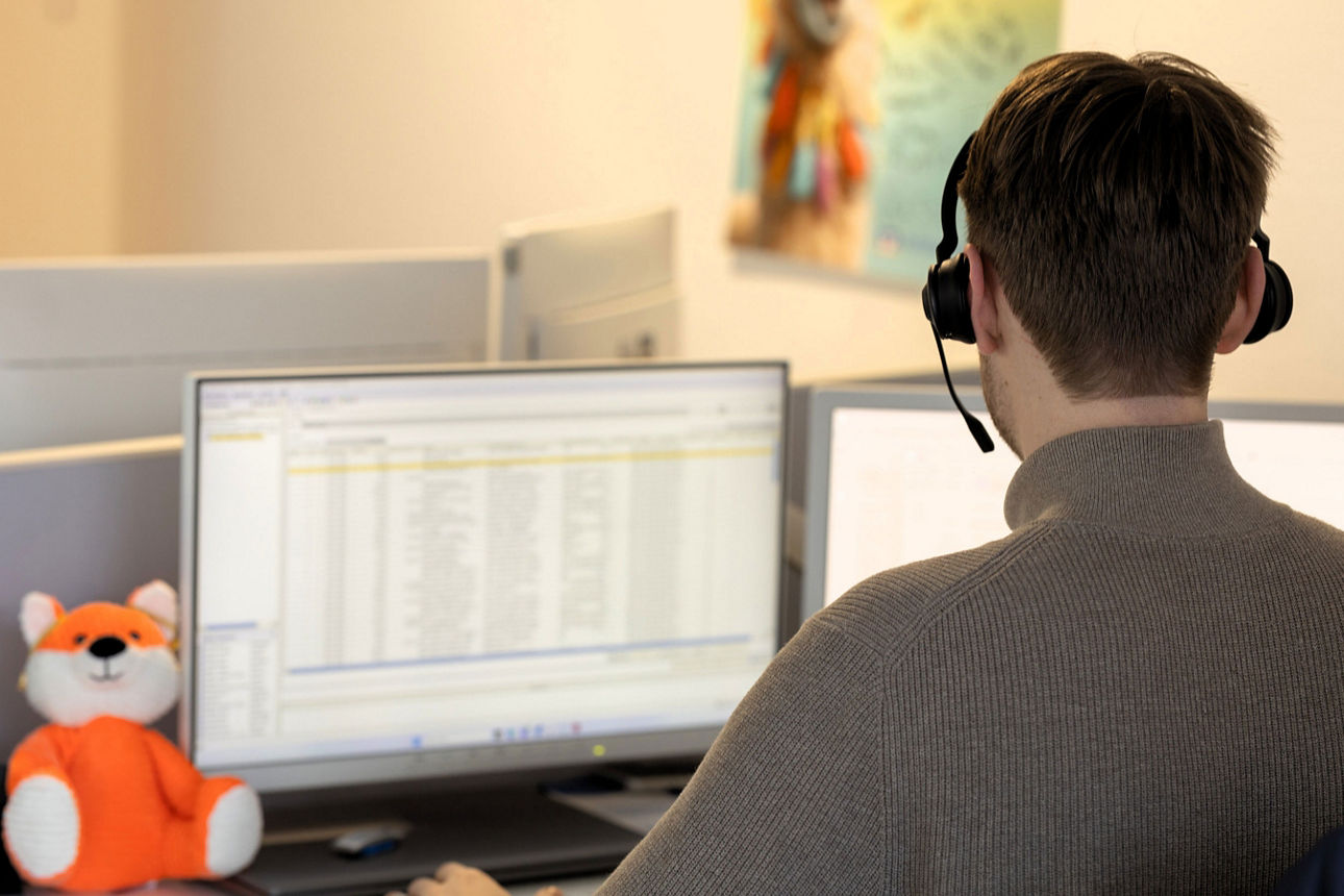 Ein Mitarbeiter der Volksbank Baumberge eG arbeitet mit Headset am PC, neben ihm sitzt ein kleiner Plüschfuchs auf dem Schreibtisch.