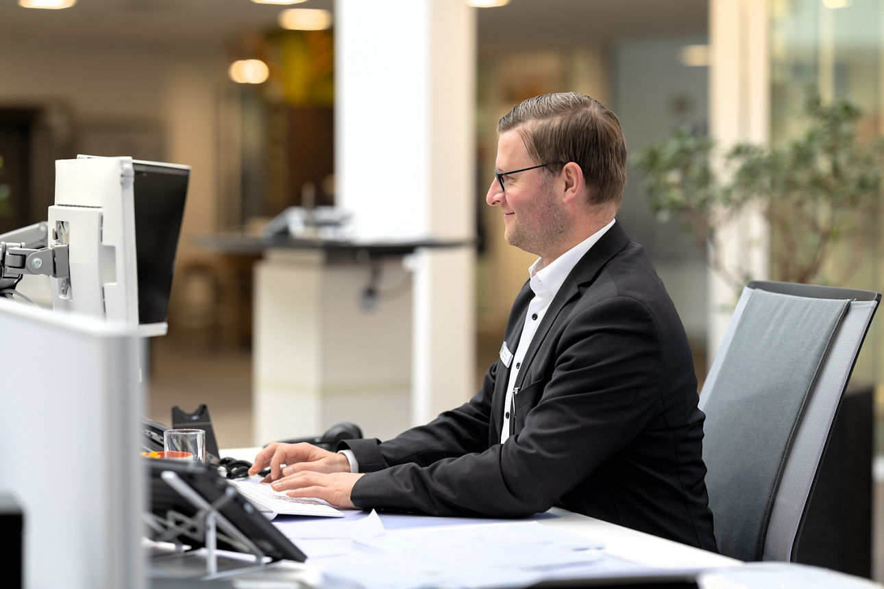 Eine männliche Person sitzt an einem Schreibtisch in einem modernen Büro und arbeitet an mehreren Monitoren.