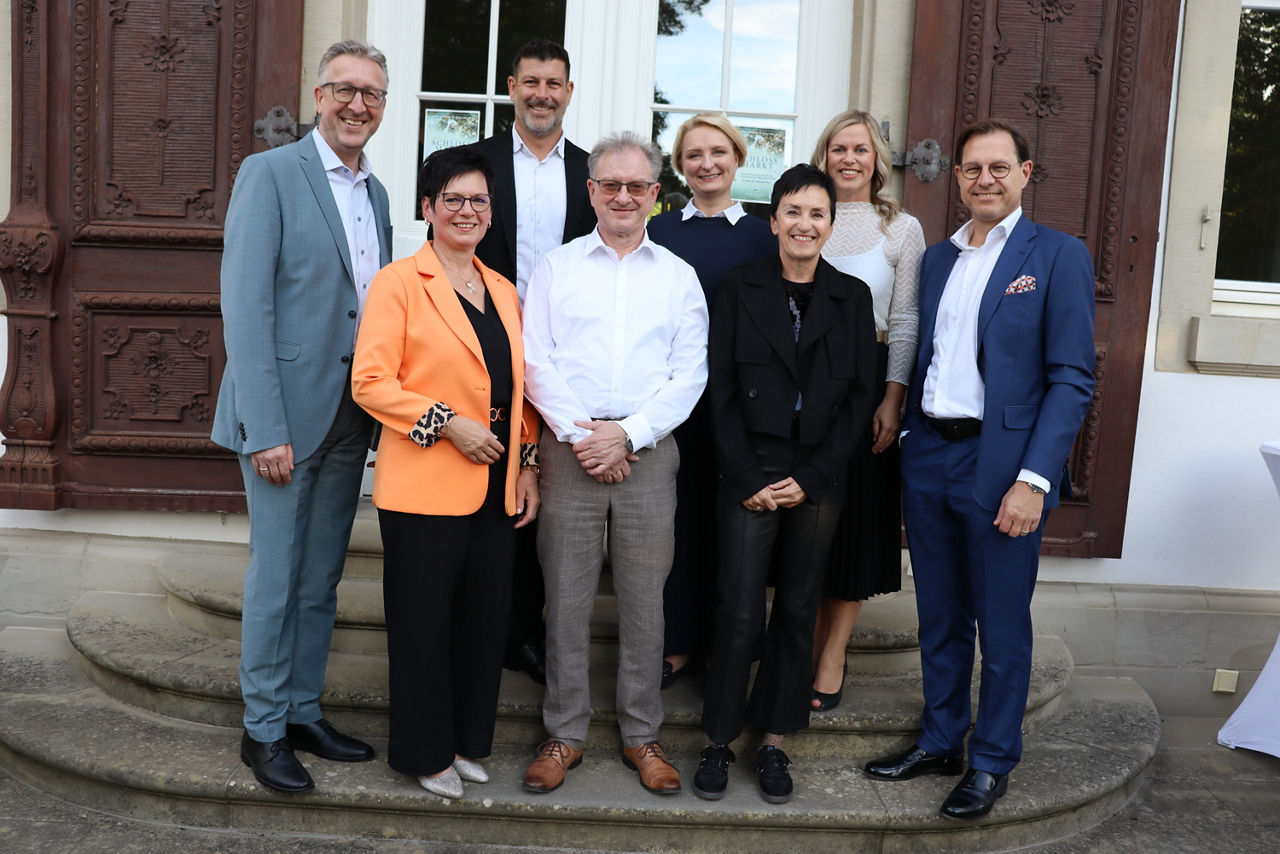 Auf dem Bild sind die Jubilare und Ihre Führungskräfte mit dem Vorstandsvorsitzenden und der Personalleiterin der Bank zu sehen. Sie stehen im Freien auf einer Treppe und lächeln in die Kamera.