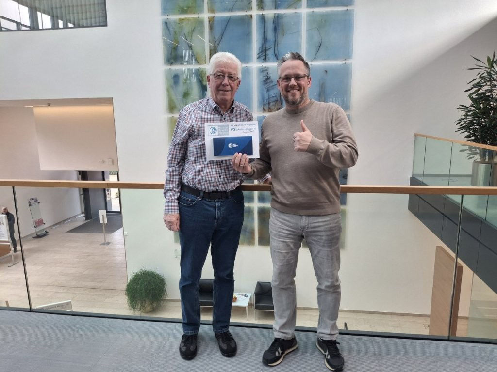 Zwei Männer stehen gemeinsam in der Lobby eines modernen Gebäudes und halten ein Zertifikat mit Markenlogo in der Hand.