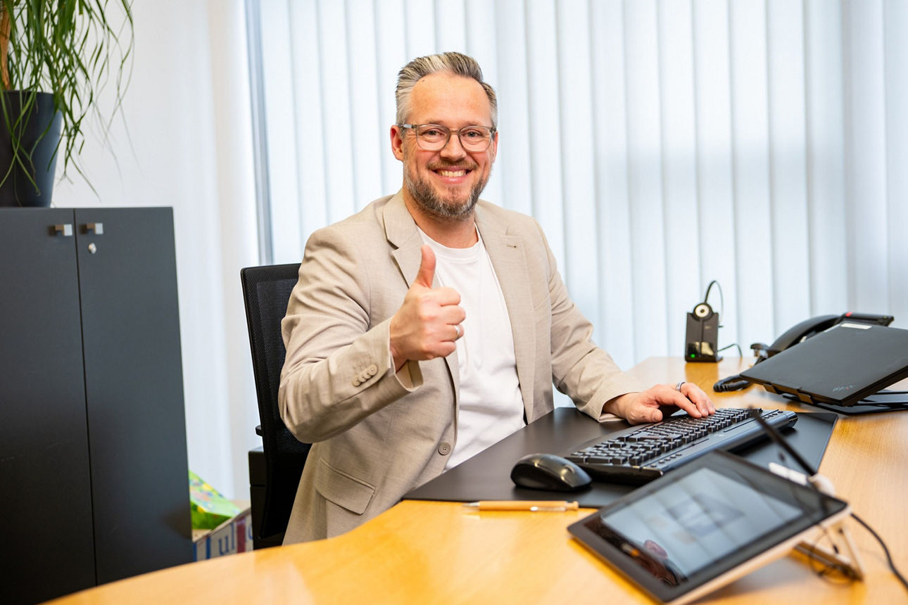 Ein Mann sitzt mit Daumen hoch lächelnd am Schreibtisch in einer Bank