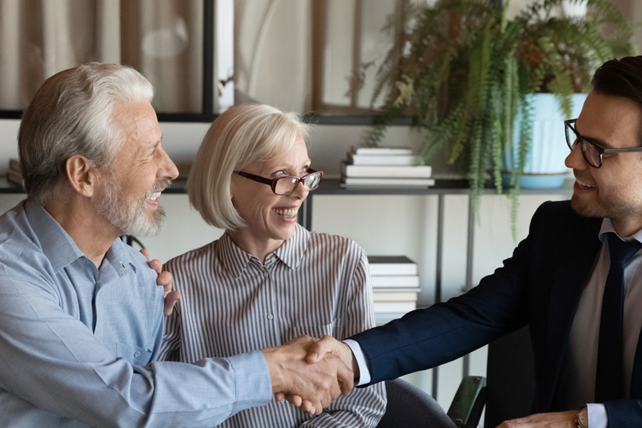 Mann im Anzug schüttelt einem anderen Mann die Hand, neben ihm sitzt eine Dame