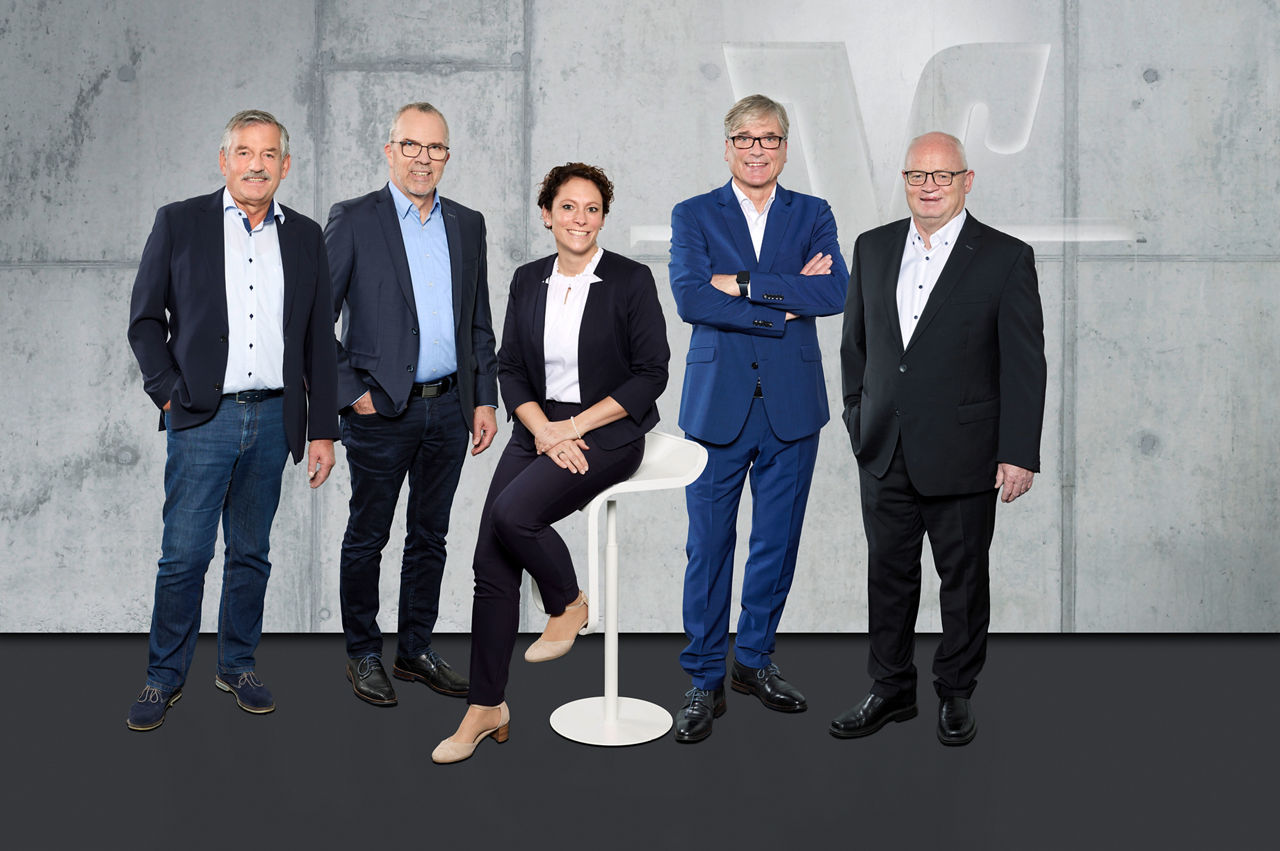 Es sind vier Männer und eine Frau in Business Kleidung zu sehen. Sie lächeln in die Kamera.