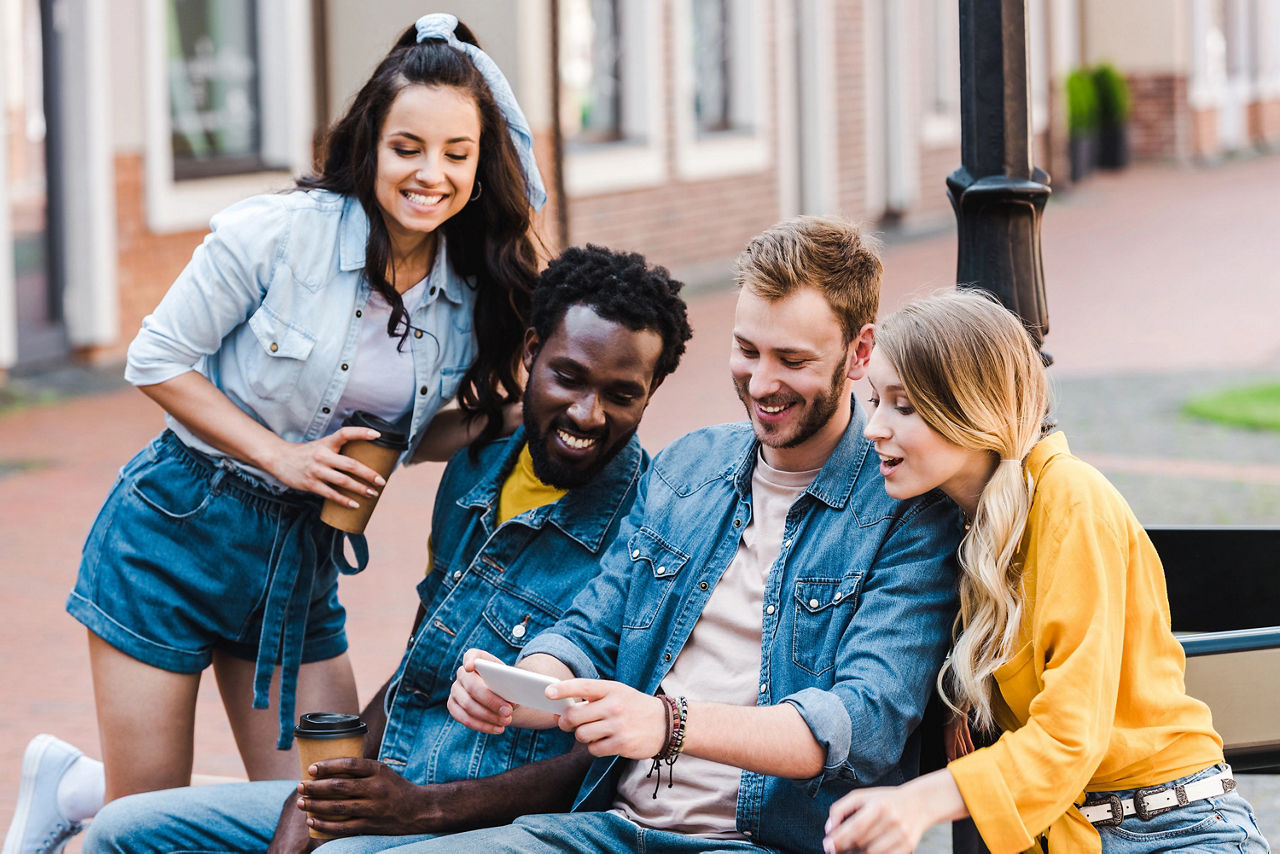 Vier Personen sitzten im Freien auf einer Bank und schauen sich freudig etwas auf einem Smartphone an.