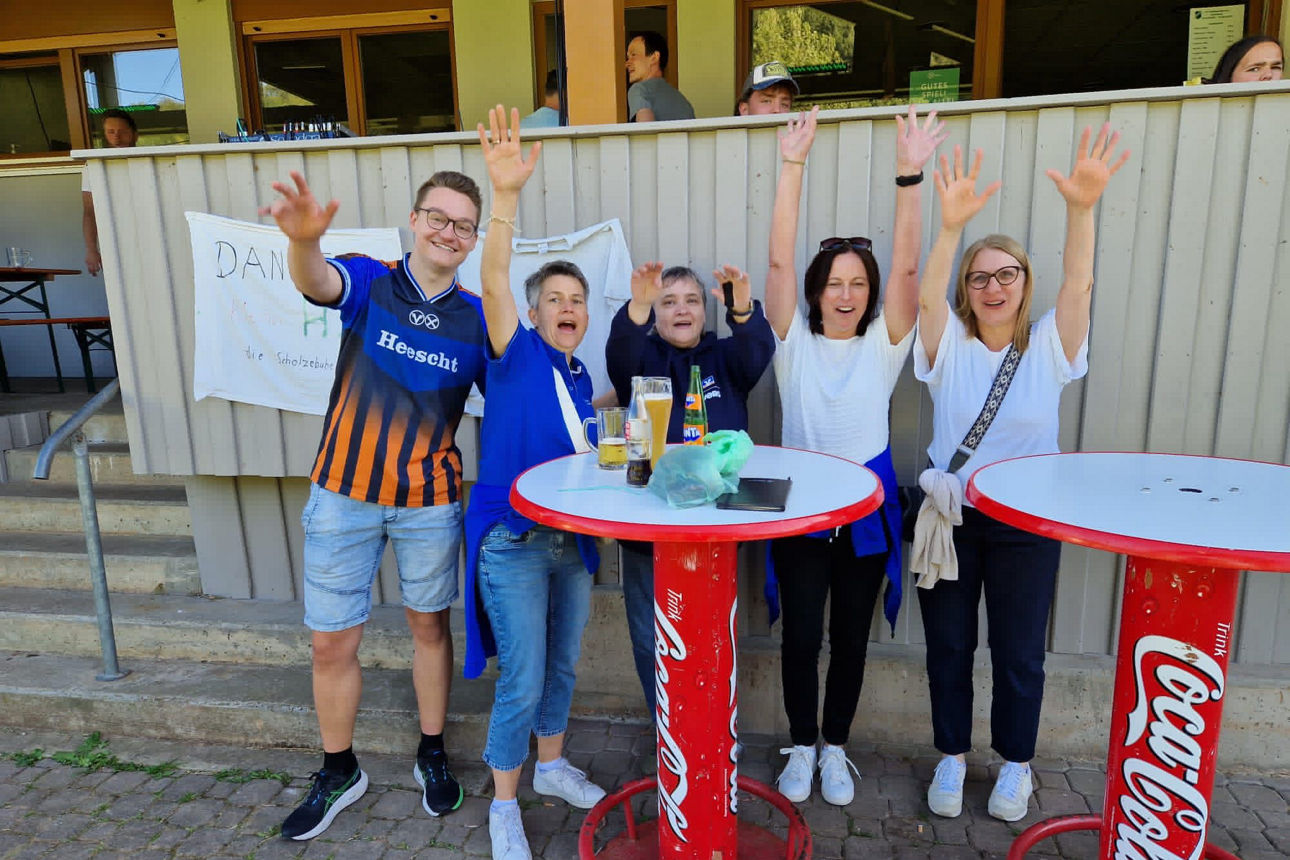 Fünf Fans an Stehtischen heben die Arme und jubeln; Getränke auf den Tischen vor dem Vereinsheim.