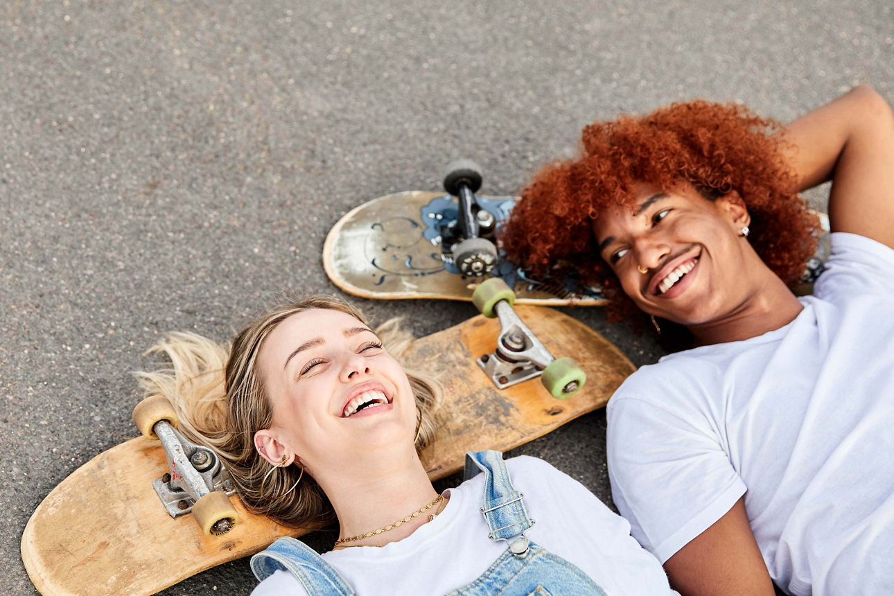 Ein junges Mädchen und ein Junge liegen lachend auf der Straße und benutzen ihre Skateboards als Kopfkissen