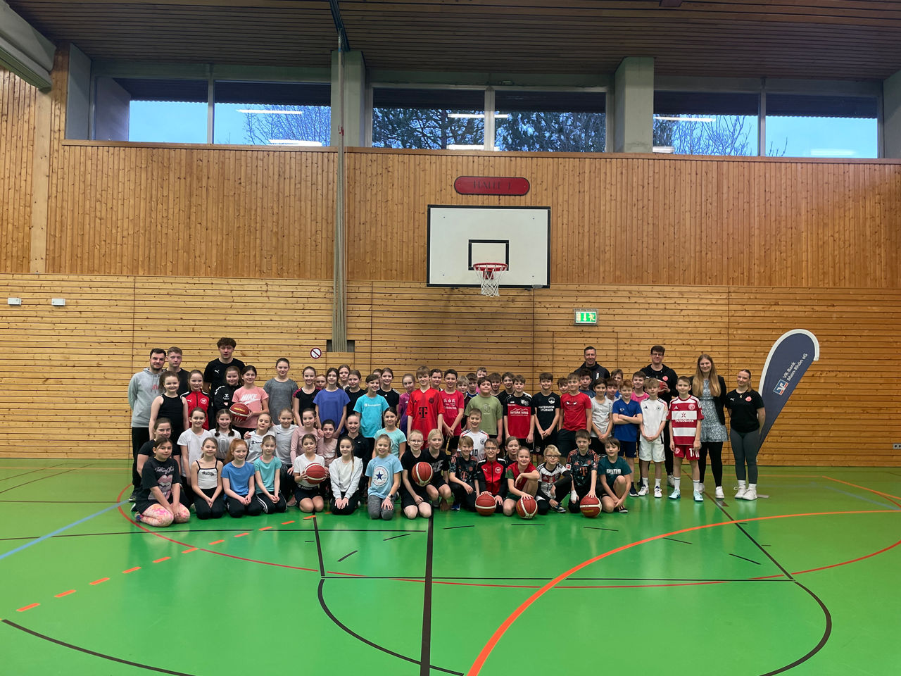 Das Bild zeigt eine große Gruppe von Kindern und Jugendlichen in einer Sporthalle, viele mit Basketbällen in den Händen. Sie 