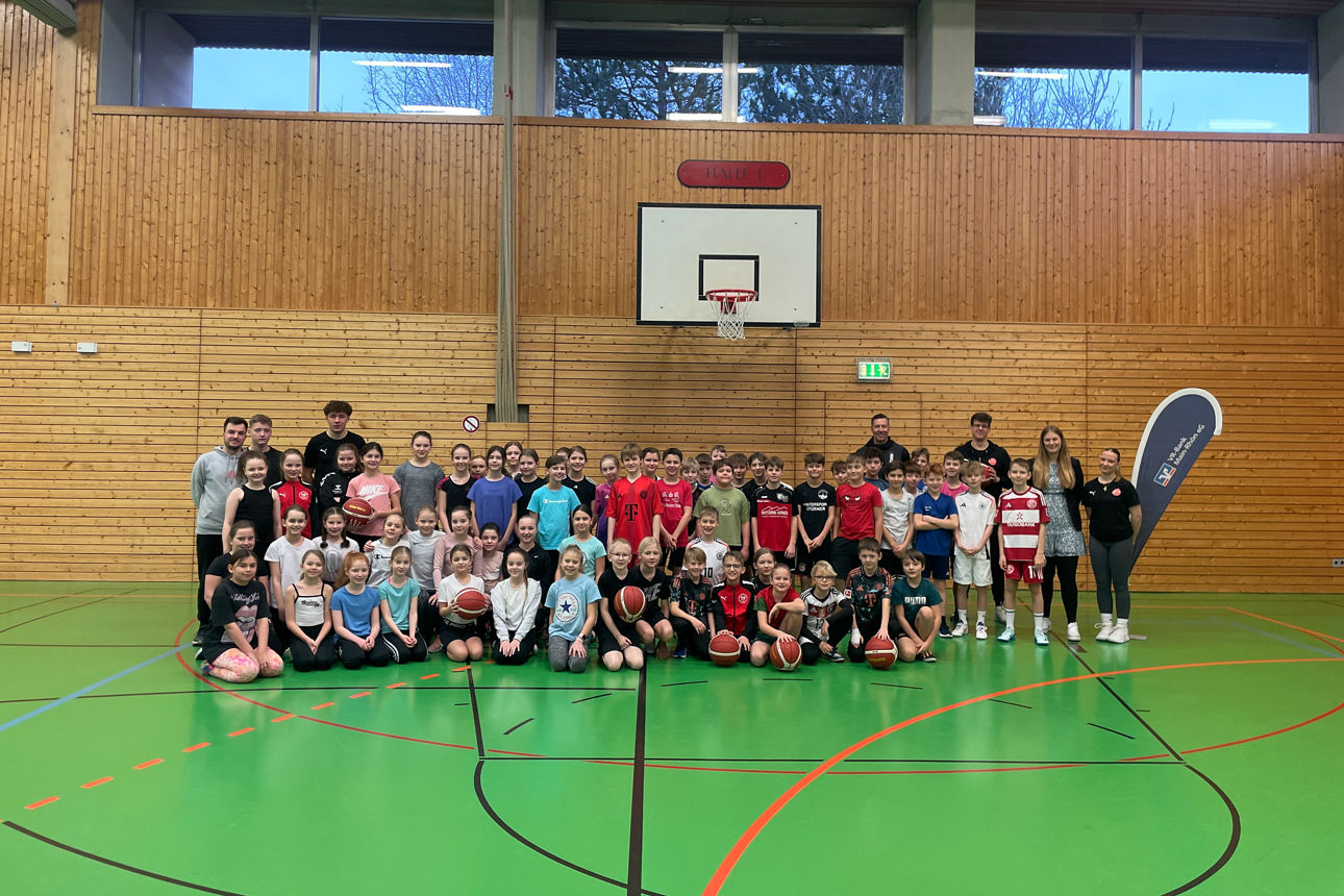 Das Bild zeigt eine große Gruppe von Kindern und Jugendlichen in einer Sporthalle, viele mit Basketbällen in den Händen. Sie 