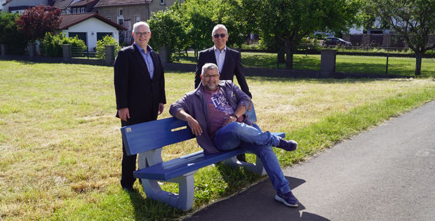 Drei Männer stehen und sitzen auf einer blauen Bank in einer grasbewachsenen Parklandschaft