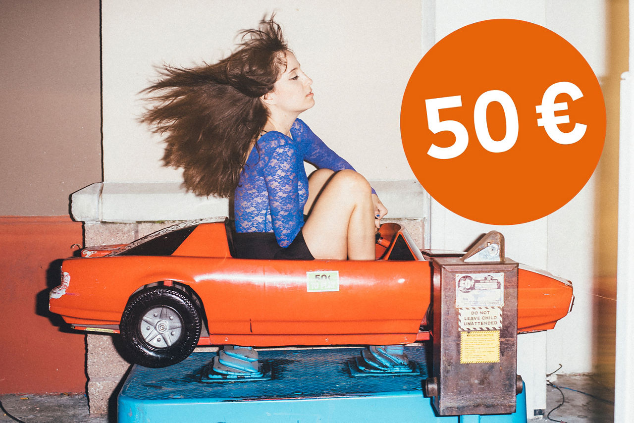 Junge Frau sitzt im Spielzeugauto, die Haare wehen nach hinten und ein Störer mit 50 €