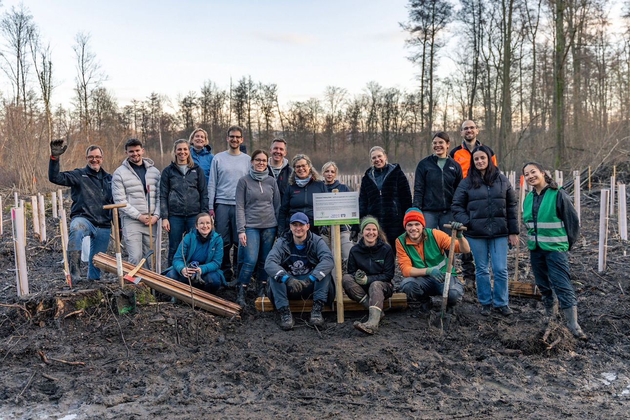 Gruppe von Helfenden steht im neu bepflanzten Waldareal vor jungen Bäumen und einer Hinweistafel