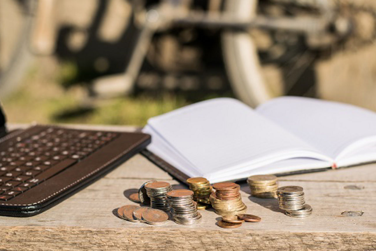 Black tablet with keyboards, an open notebook with white pages and a few stacks of coins on a wooden table. Outdoors photo under the direct light of sun.