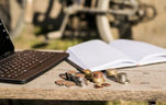 Black tablet with keyboards, an open notebook with white pages and a few stacks of coins on a wooden table. Outdoors photo under the direct light of sun.