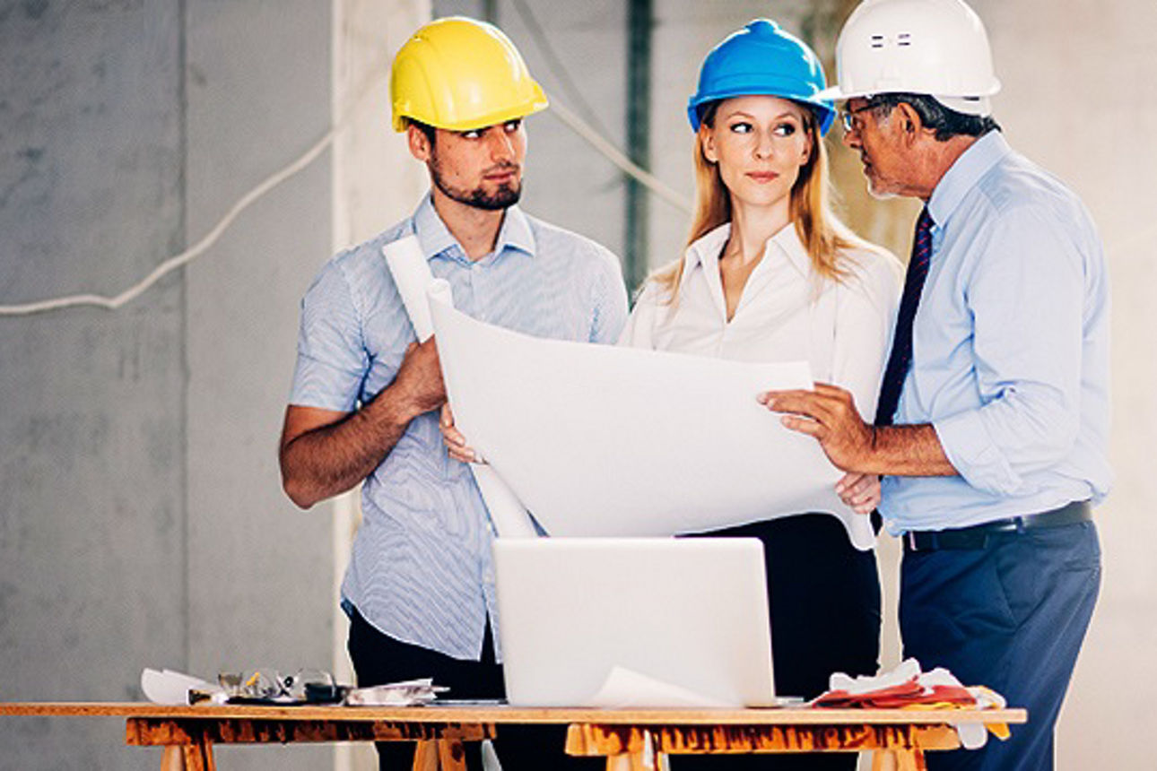 Drei Personen mit Schutzhelmen prüfen gemeinsam Baupläne auf einer Baustelle