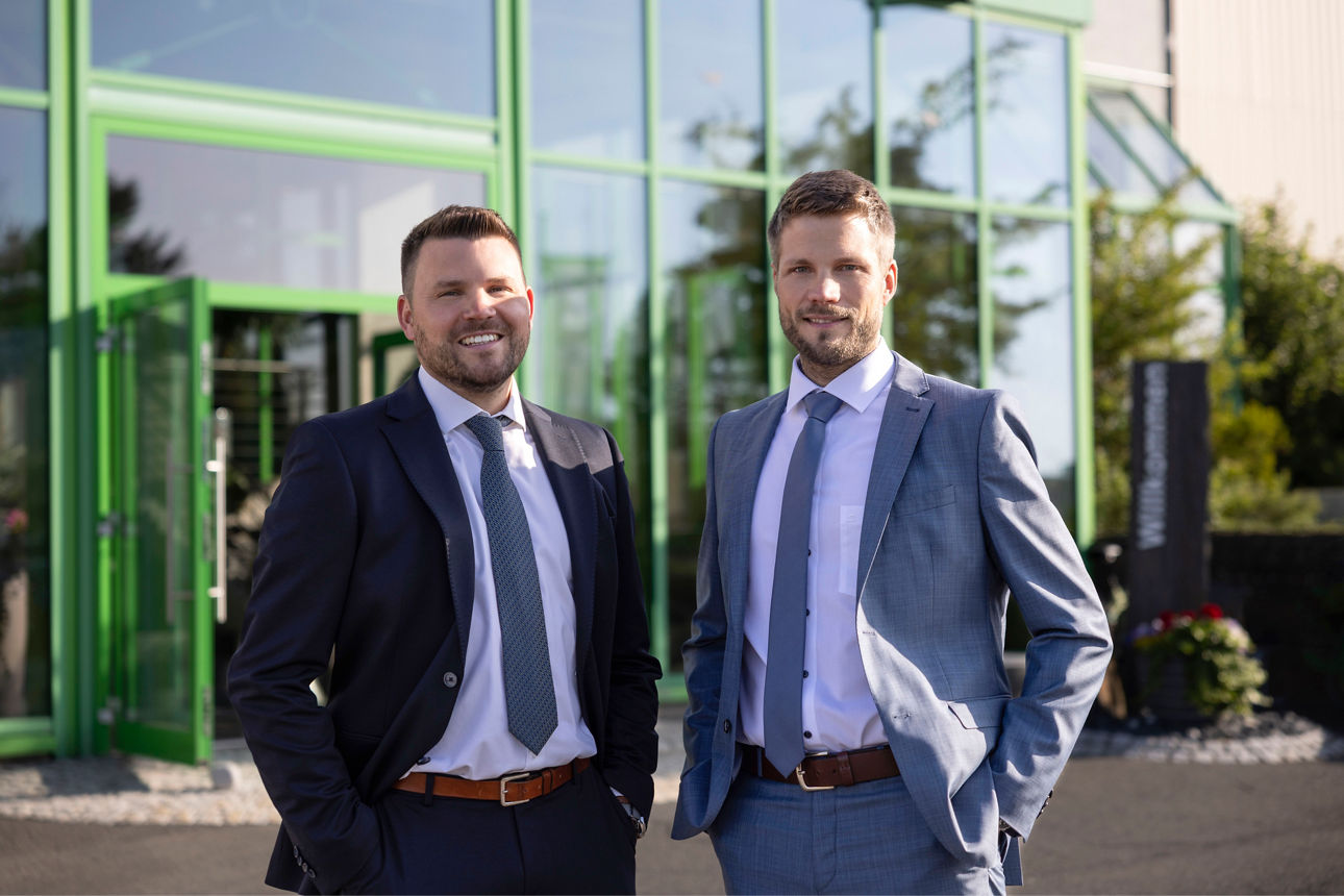 Zwei Geschäftsleute stehen selbstbewusst vor einem modernen grünen Gebäude.