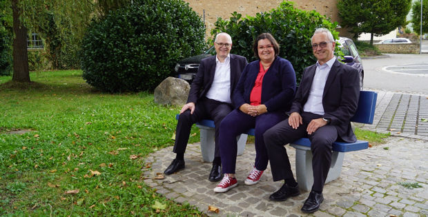 Drei Personen, zwei Männer und eine Frau, sitzen draußen auf einer blauen Bank.