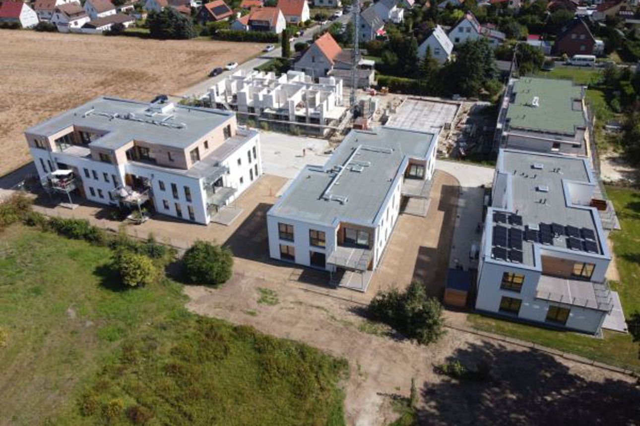 Luftaufnahme mehrerer moderner Flachdachgebäude in einer Neubausiedlung, umgeben von Feldern und Einfamilienhäusern.