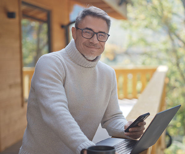 Ältere Mann mit Brille auf dem Balkon mit Handy und Laptop