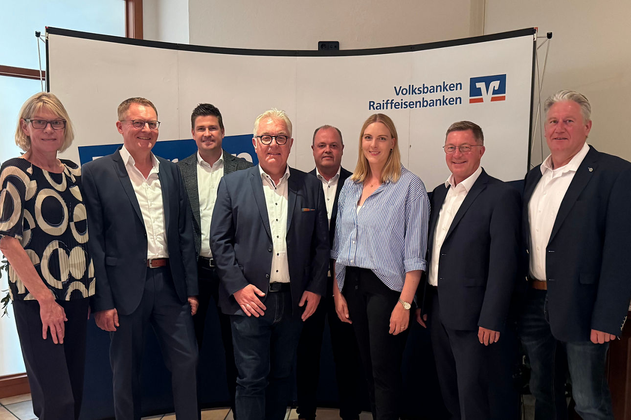 Eine Gruppe von acht Personen, die vor einem Hintergrund mit dem Logo der Volksbanken Raiffeisenbanken posieren.