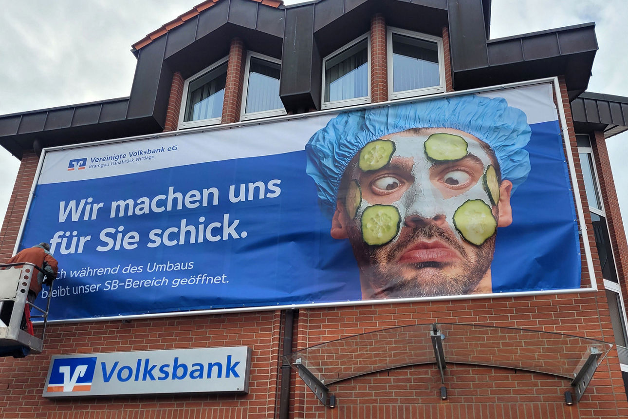 An einem Backsteingebäude wird ein großes Plakat aufgehängt, auf dem steht "Wir machen uns für Sie schick."