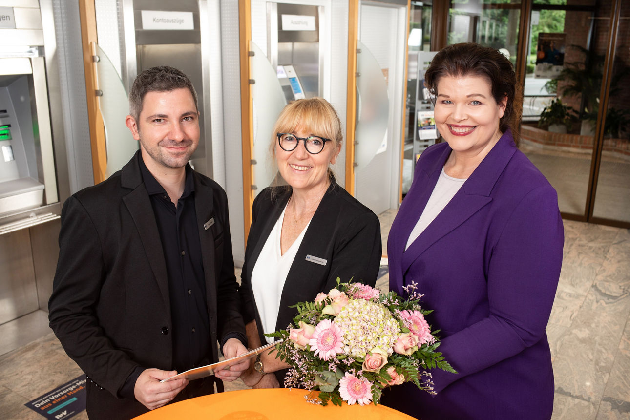 Ein Mann und zwei Frauen stehen im SB-Bereich einer Bank. Sie halten Blumen und einen Brief in der Hand.