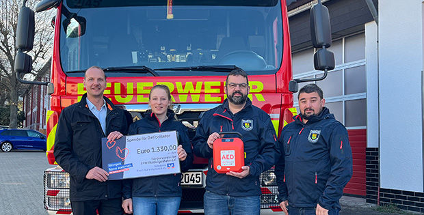 Vier Personen stehen vor einem Feuerwehrwagen, zwei halten einen Spendenscheck in der Hand.