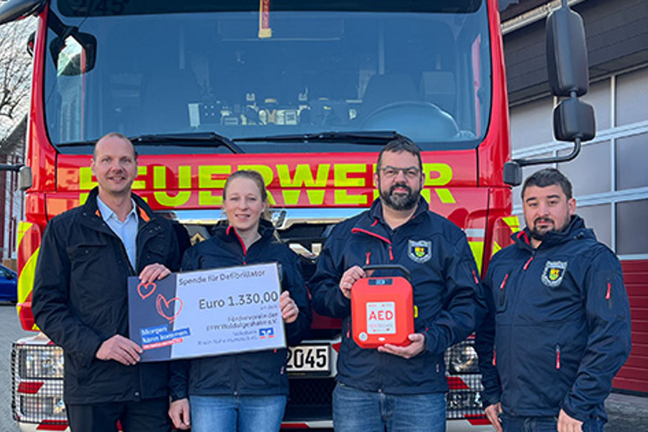 Vier Personen stehen vor einem Feuerwehrwagen, zwei halten einen Spendenscheck in der Hand.