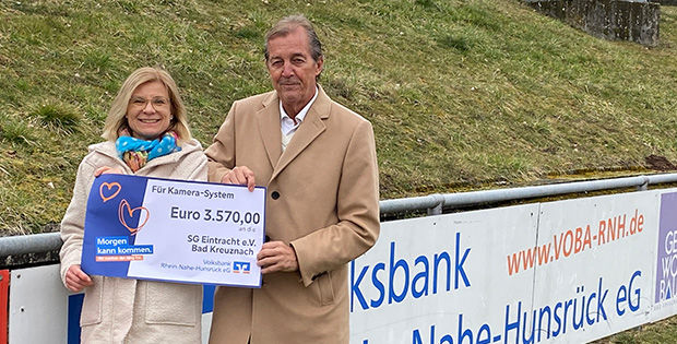 Ein Mann und eine Frau auf einem Sportplatz, sie tragen einen Spendenscheck in der Hand.