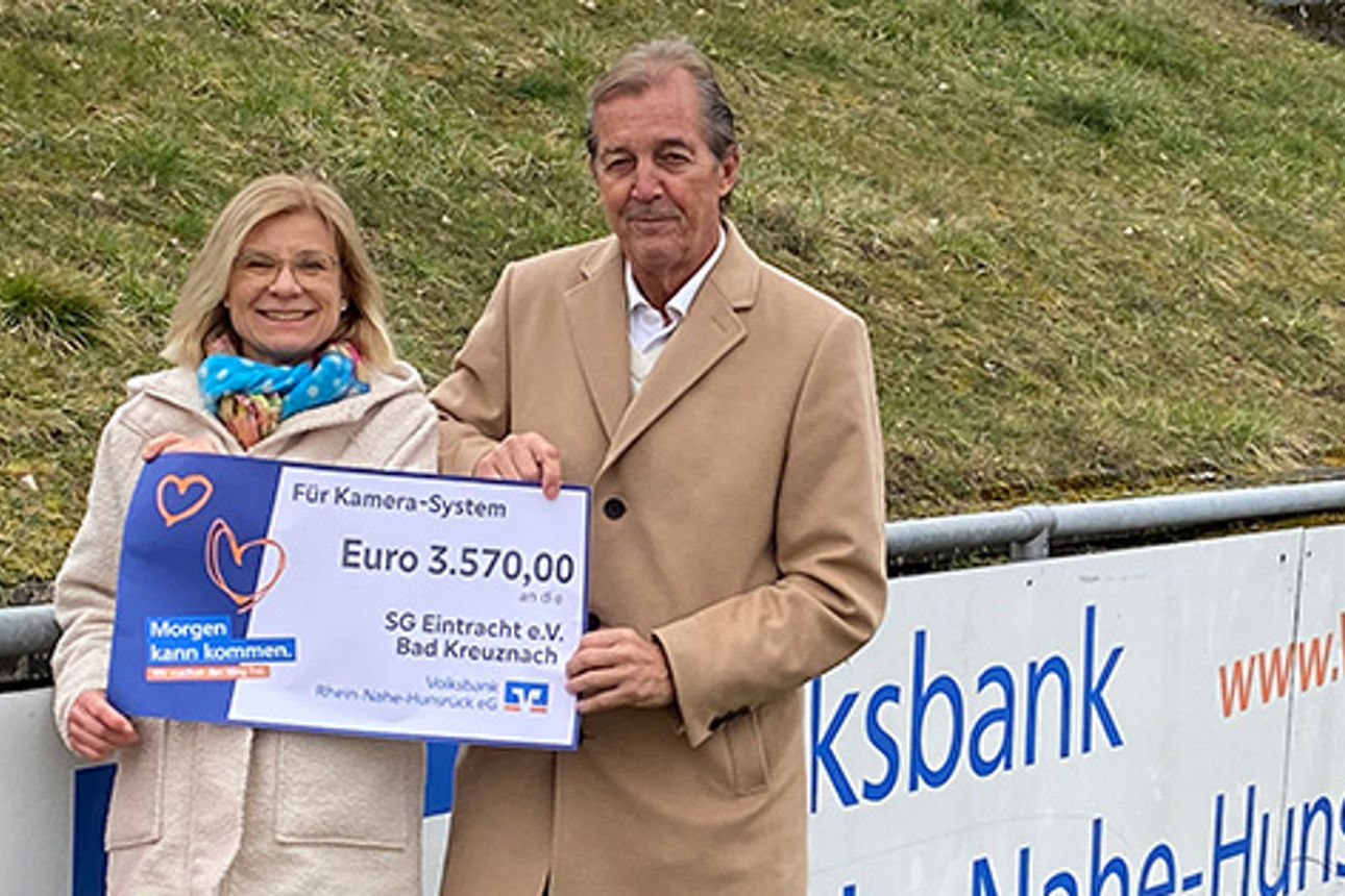 Ein Mann und eine Frau auf einem Sportplatz, sie tragen einen Spendenscheck in der Hand.