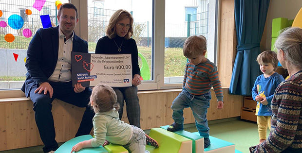 Zwei Erwachsene sitzen in einer Kita und halten einen Spendenscheck in den Händen. Um sie herum spielen Kinder.
