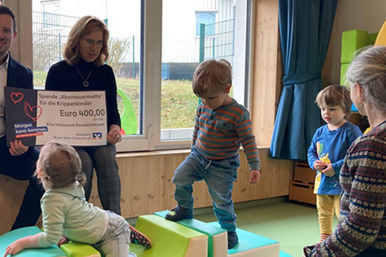 Zwei Erwachsene sitzen in einer Kita und halten einen Spendenscheck in den Händen. Um sie herum spielen Kinder.