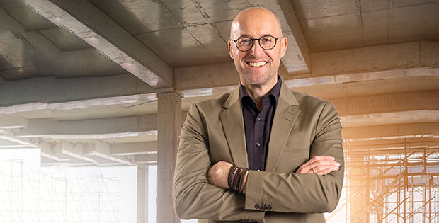 Axel Müller steht in einem Rohbau. Er trägt eine Brille und einen braunen Anzug mit schwarzem Hemd.