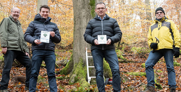 Vier männliche Personen vor einem Baum. Zwei halten eine Auszeichnung in Händen. 