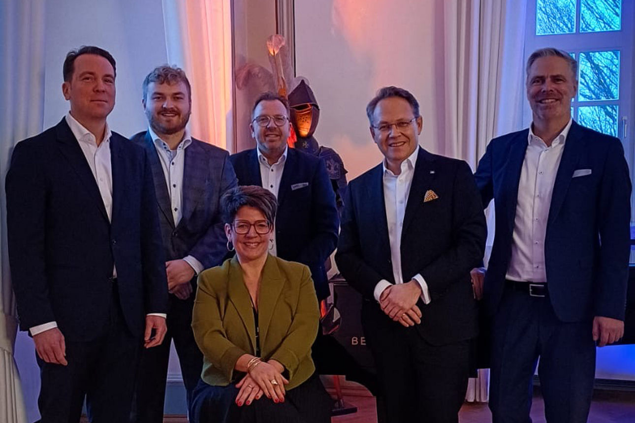 Eine Gruppe von Personen in formeller Kleidung steht um einen Flügel in einem eleganten Raum mit großen Fenstern und Holzbode