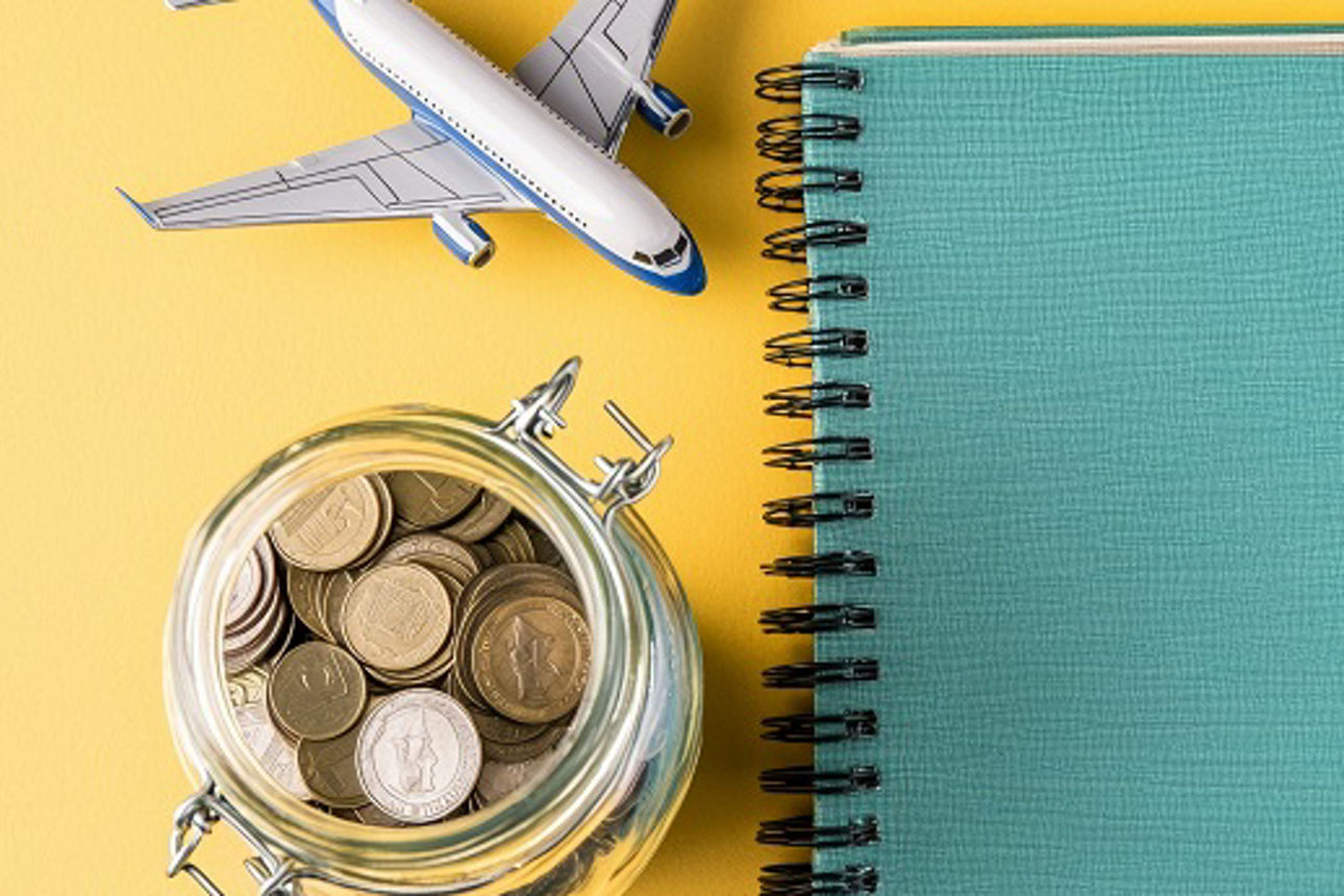 Mit fremdländischen Münzen gefülltes Glas neben Notizblock und symbolisch dazugestelltem Modellflugzeug