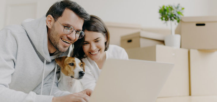 Paar mit Hund vor Laptop vor Umzugskartons im Hintergrund