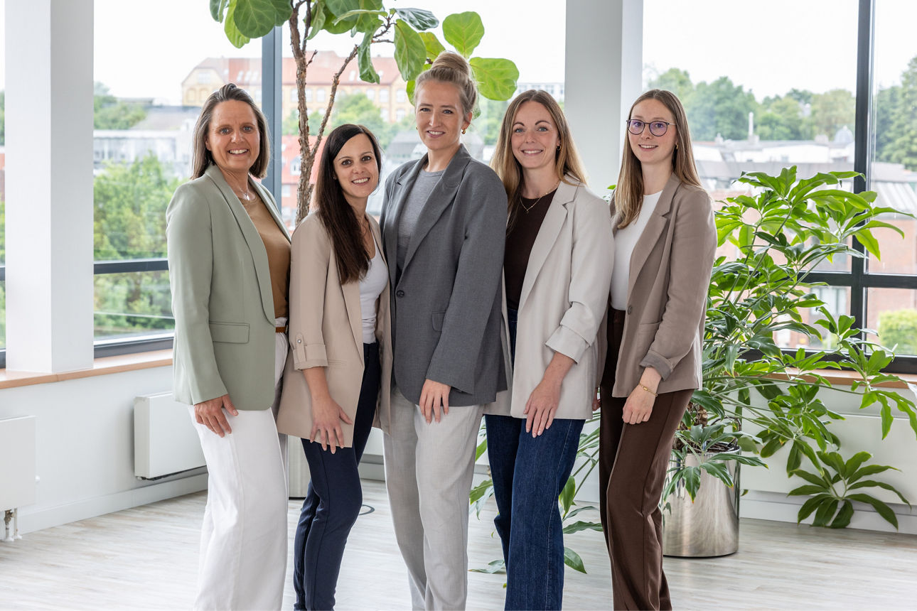 Fünf Frauen stehen nebeneinander in einem hellen Raum mit großen Fenstern. 