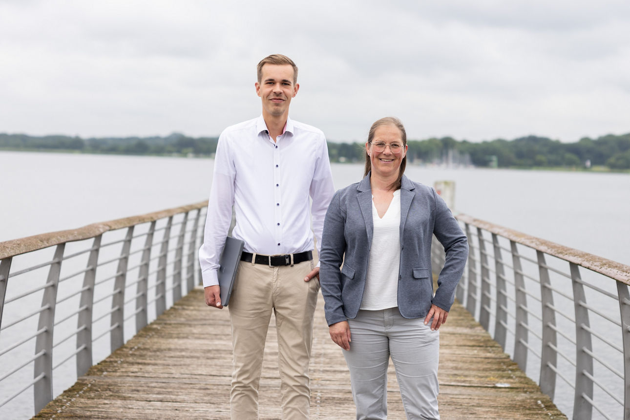 Zwei Personen in Businesskleidung stehen auf einem Steg mit Geländer vor einem Gewässer und bewölktem Himmel