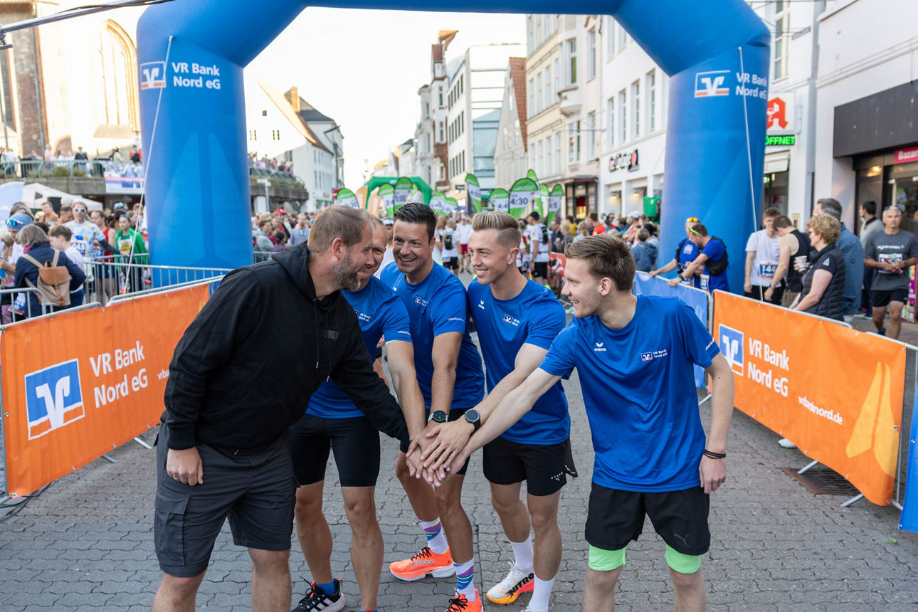 Gruppe von Läufern in blauen Shirts und einem Mann in schwarzer Sportkleidung, die vor einem Startbogen mit dem Schriftzug VR Bank Nord eG die Hände zusammenlegen.