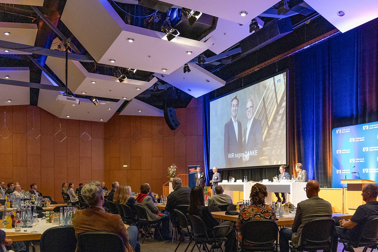 Man sieht das innere der Stadthalle Gladbeck, inklusive der Bühne und der großen Projektorleinwand. Auf der Bühne stehen und sitzen vier Personen. Bei zwei dieser Personen handelt es sich um den Vorstand der Volksbank Ruhr Mitte, die anderen beiden Personen stammen aus dem Aufsichtsrat. Im Vordergrund sind viele sitzende Zuhörer zu sehen. Es handelt sich um eine Momentaufnahme der Vertreterversammlung der Volksbank Ruhr Mitte.